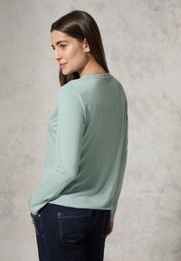 Helles grünes Langarmoberteil mit Rundhalsausschnitt, aus weichem Stoff mit dezenter Textur. Wird mit dunkelblauen Jeans getragen, Rückansicht.