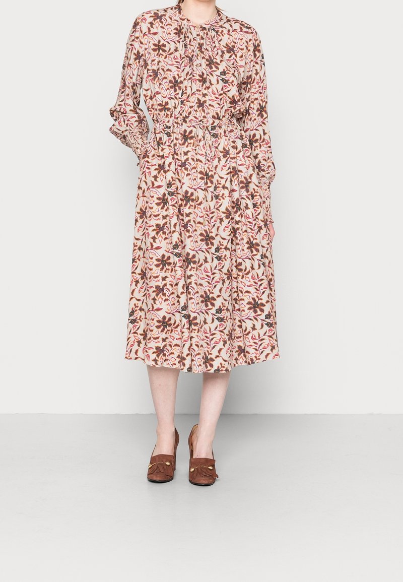 Femme portant une robe midi à manches longues avec un motif floral rose, marron et rouge, associée à des mocassins à talons marron, debout sur un sol gris.