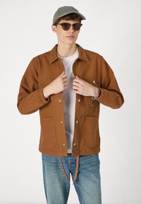 Bruine, knopen-jack met lange mouwen, elastische manchetten, voorzakken en een trekkoord onderaan. Draag het over een wit shirt met blauwe jeans.