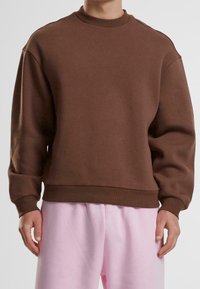 Felpa marrone con colletto rotondo, spalle abbassate e polsini a costine. Tessuto semplice, abbinata a pantaloni rosa chiaro.