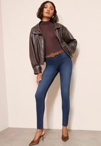 Chaqueta bomber de cuero marrón, cuello de tortuga bronce ajustado, jeans skinny azul oscuro, cinturón marrón y mule de tacón alto marrones sobre un fondo neutro.