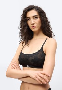 Νεαρή γυναίκα με σγουρά μαλλιά, φορώντας μαύρο φλοράλ δαντελένιο μπραλέτο, με σταυρωμένα χέρια, κοιτάζει με αυτοπεποίθηση την κάμερα σε απλό φόντο.