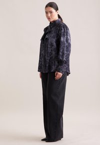 Dunkelgrau Bluse mit Leopardenmuster, hohem Kragen und Schleifendetail. Verfügt über lange Ärmel mit gerafften Manschetten. Kombiniert mit schwarzen, weit geschnittenen Hosen.
