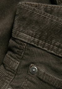 Brązowa tkanina sztruksowa z fakturowanym prążkowanym wzorem, z widocznym metalowym guzikiem oznaczonym napisem "DENIM" oraz starannym stebnowaniem.