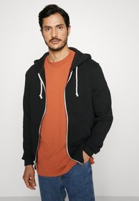 Zwarte zip-up hoodie met witte drawstrings, gedragen over een oranje t-shirt, gecombineerd met blauwe jeans. De stof lijkt zacht en casual.