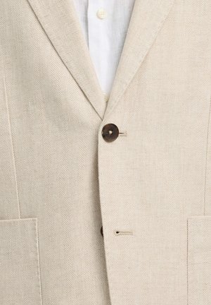 Bež bež s vzorcem ribje kosti, zapet preko bele srajce z vidnimi kremnimi gumbi in z odprtim ovratnikom.