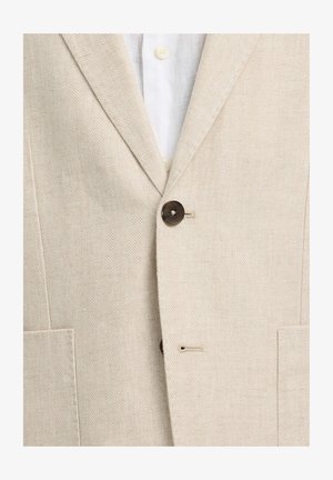 Bež bež s vzorcem ribje kosti, zapet preko bele srajce z vidnimi kremnimi gumbi in z odprtim ovratnikom.