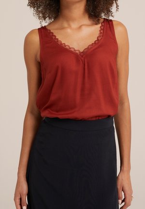 Vrouw draagt een mouwloze, roestkleurige V-hals top met kant, ingestopt in een zwarte hoge taille rok, staand tegen een neutrale achtergrond.
