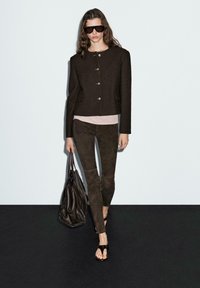 Brown wool-blend jacket with gold button closures, worn over a light pink top and paired with dark brown suede pants and black sandals.