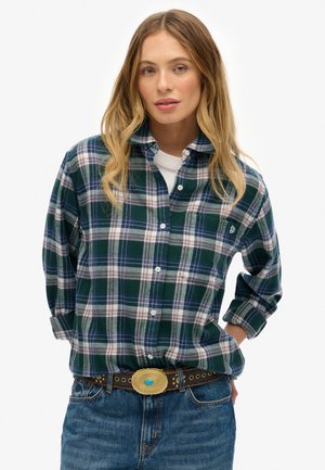 Green and navy plaid flannel shirt with a button-down collar, worn over a white shirt, paired with denim and a gold-accented belt.