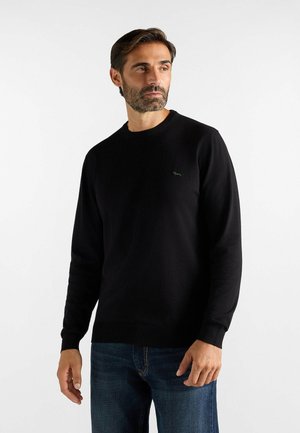 Uomo con la barba che indossa un maglione nero a maniche lunghe e jeans blu, guarda leggermente a destra, in piedi davanti a uno sfondo bianco semplice.