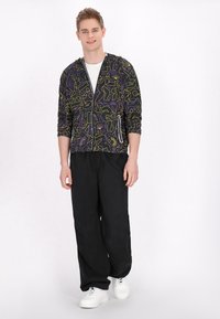 Giacca a zip nera con cappuccio e motivi astratti multicolore, abbinata a pantaloni neri larghi e sneakers bianche. Sotto indossa una semplice maglietta bianca.