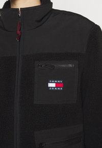 Czarna polarowa kurtka z zamkiem błyskawicznym, kieszenią na suwak, tekstylnymi detalami oraz naszywką z logotypem "TOMMY JEANS" w kolorach niebieskim, czerwonym i białym.