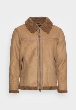 Veste en shearling beige avec fermeture éclair frontale, deux poches latérales zippées, et un col ainsi que des poignets en fausse fourrure douce.