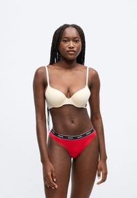Béžová podšitá podprsenka s tenkými ramienkami a červené bikini nohavičky s čiernym pásom s logom „HUGO“, na jednoduchom pozadí.