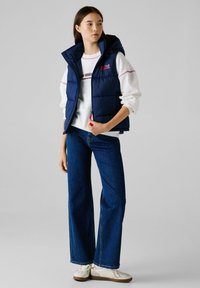 Μπέζ αμάνικο μπουφάν του ναυτικού με κουκούλα, φερμουάρ στο μπροστινό μέρος και δύο τσέπες, φορεμένο πάνω από έναν λευκό φούτερ. Σε συνδυασμό με φαρδιά μπλε τζιν και λευκά αθλητικά παπούτσια.