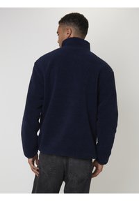 Man in een marineblauwe fleecejack met hoge kraag, staande met zijn rug naar de camera tegen een effen achtergrond.