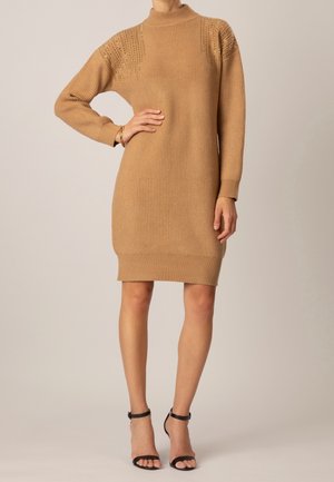 Kvinde iført en knælængde sweaterkjole i kamelfarvet strik med lange ærmer og sorte høje hæle, stående med den ene hånd på hoften.