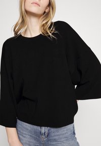 Czarny dziergany sweter z oversize'owymi rękawami i okrągłym dekoltem, zestawiony z jasnoniebieskimi dżinsami. Charakteryzuje się luźnym krojem i teksturowaną tkaniną.