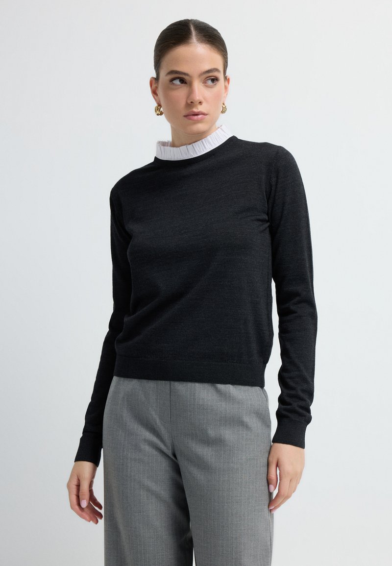 Zwarte trui met lange mouwen en een ronde hals, gelaagd over een witte, gerimpelde kraag. Draagt met grijze pantalon met streepjes.