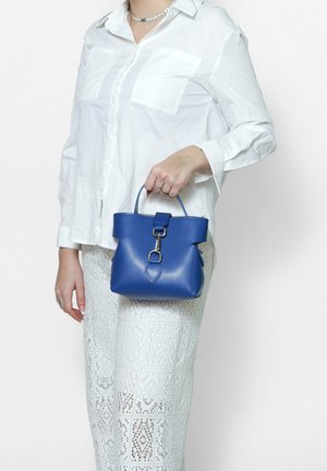 Borsa in pelle blu con forma strutturata, manico corto, tracolla frontale e hardware in metallo. Indossata con una camicia bianca e pantaloni con motivo in pizzo.