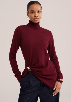 Femme portant un pull à col roulé rouge foncé et un pantalon bleu foncé, debout avec une main dans la poche devant un fond uni.