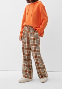 Oranje hoodie met een ontspannen pasvorm, gecombineerd met wijde ruitbroek in bruin, crème en oranje tinten, aangevuld met witte sneakers.