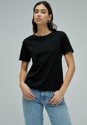 Mujer con cabello largo y oscuro, vestida con una camiseta negra lisa de manga corta y jeans azul claro, de pie con las manos cerca de los bolsillos.
