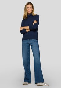 Marineblauer Rollkragenpullover mit geripptem Muster und Knopfdetails an den Ärmeln, kombiniert mit ausgestellten blauen Jeans und silbernen Schuhen.