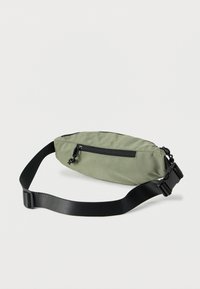 Πράσινη τσάντα μέσης από νάιλον με μαύρες λεπτομέρειες, λεπτή σχεδίαση, ρυθμιζόμενο ιμάντα, μπροστινή τσέπη με φερμουάρ και ανθεκτικό εξοπλισμό. Λεπτομέρεια με υφή στην επιφάνεια.