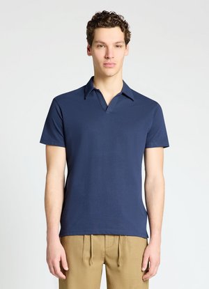 Giovane uomo che indossa una polo blu navy a maniche corte e pantaloni marrone chiaro, in piedi davanti a uno sfondo chiaro uniforme.