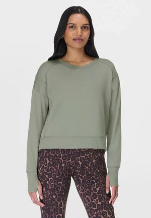 Vrouw met lang donker haar, gekleed in een lichtgroene trui met lange mouwen en bruine legging met luipaardprint, staand tegen een witte achtergrond.