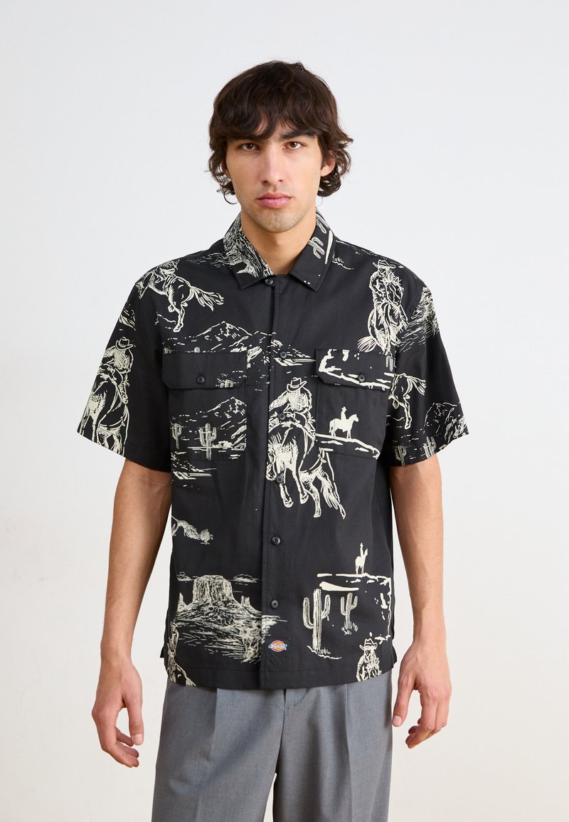 Camicia a maniche corte con bottoni, sfondo nero, presenta motivi bianchi di cowboy e deserto. Due tasche sul petto, vestibilità rilassata.