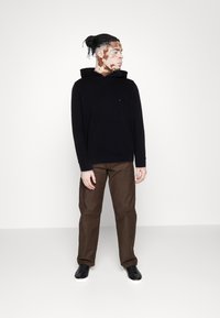 Sweatshirt à capuche noire avec une poche avant, associé à un pantalon marron et des baskets noires. Tissu lisse avec une coupe décontractée.
