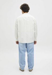 Helles beige Langarmshirt mit entspannter Passform, Rückenpasse und geknöpften Manschetten; kombiniert mit weiten blauen Jeans und hellen Schuhen.