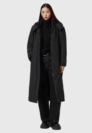 Manteau long noir en duvet avec un col montant, des fermetures éclair et des boutons-pression, et des poches latérales, porté sur un pull noir et un pantalon. Texture épaisse et lisse.