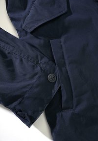 Marineblå jakke med en jevn tekstur, knappelukking og et lomme med klaff. Stoffet virker lett og litt glansfullt.