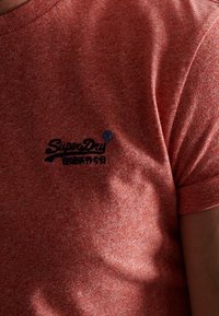 Röd melerad t-shirt med kort ärm som har en mörkblå broderad logotyp "SuperDry" på bröstet och rund halsringning.