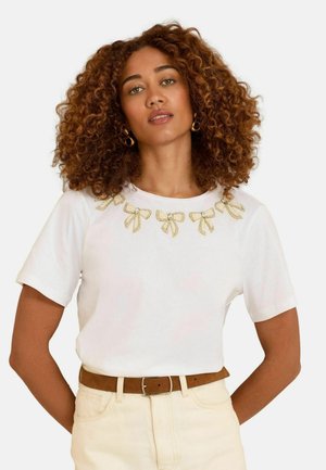 T-shirt blanc à manches courtes avec un col décoratif en nœuds dorés rehaussés de strass, associé à un pantalon de couleur claire.