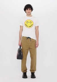 White t-shirt with a yellow circular logo featuring palm trees. Beige trousers with an adjustable drawstring and black footwear. Black and colorful handbag.