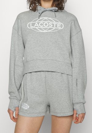 Kobieta ubrana w jasnoszary krótki kaptur i dopasowane szorty, oba z białym logo Lacoste, stojąca na jednolitym tle.