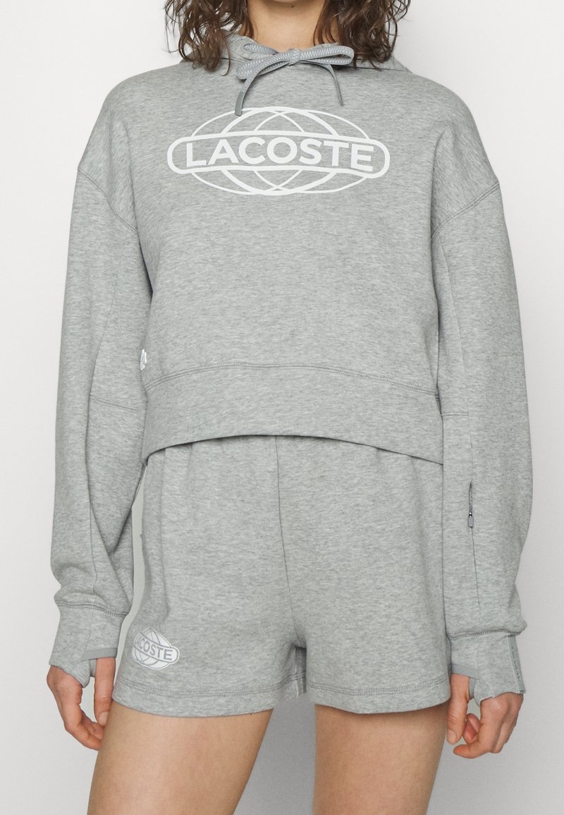 Femme portant un sweat à capuche court gris clair et un short assorti, tous deux avec le logo blanc Lacoste, debout devant un fond uni.