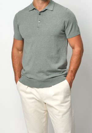 Homme portant un polo gris ajusté à manches courtes et un pantalon blanc, les mains dans les poches, sur un fond uni.