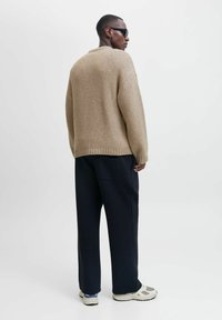 Beige Strickpullover mit Rundhalsausschnitt, kombiniert mit lockeren schwarzen Hosen und Sneakers. Der Pullover hat ein strukturiertes, grobmaschiges Design.