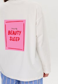 Cremefarbenes Oversized-Sweatshirt mit einer pinken Grafik auf dem Rücken. Der Text lautet: "Ich brauche meinen SCHÖNHEITSSCHLAF" in fetten roten Buchstaben. Weicher Stoff.