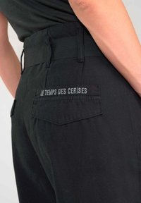 Zwarte katoenen broek met een elastische tailleband, voorzien van een achterzak met "LE TEMPS DES CERISES" in witte letters gestikt. Gladde textuur.