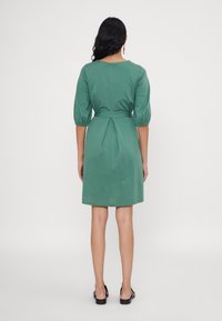 Frau mit langen dunklen Haaren, die ein knielanges grünes Kleid mit drei Viertelärmeln und schwarzen flachen Schuhen trägt, steht mit dem Rücken zu uns.