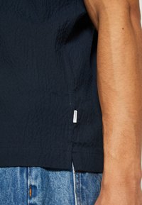 Camisa texturizada de color marino con dobladillo redondeado, abertura lateral y una pequeña etiqueta en la costura. Usada con pantalones de mezclilla azul claro.