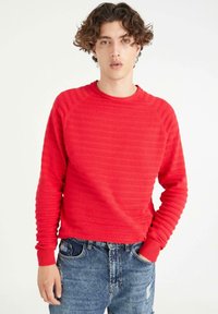 Jersey rojo texturizado con puños y cuello acanalados, que presenta rayas horizontales. Combinado con jeans de mezclilla desgastados.