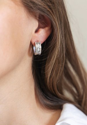 Gros plan sur l'oreille d'une femme avec des cheveux brun moyen portant des boucles d'oreilles en argent en forme de créole ornées de pierres précieuses transparentes.